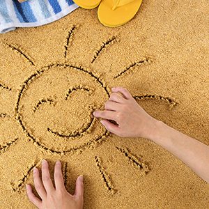 Child drawing a smiley sun in sand with towel, seashells and flip flops